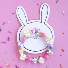 Load image into Gallery viewer, A colorful Easter-themed bracelet with bunny and egg charms, displayed on a bunny-shaped card against a pink background.