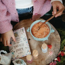 Load image into Gallery viewer, A cozy tabletop scene with a person in a pink fuzzy cardigan holding a book and stirring a small bowl of orange liquid with a wooden spoon. The setup includes small jars, moss, flowers, and a rustic wooden surface.