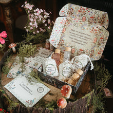Load image into Gallery viewer, A styled product display of a woodland-themed gift box set on a tree stump. The open box contains several small bottles, wrapped items, and printed cards with whimsical illustrations of fairies and mushrooms, surrounded by pink flowers, greenery, and decorative props.