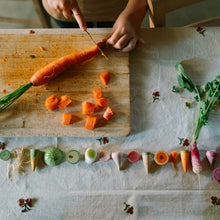 Load image into Gallery viewer, Person cutting a carrot on a wooden cutting board with various vegetables arranged below.
