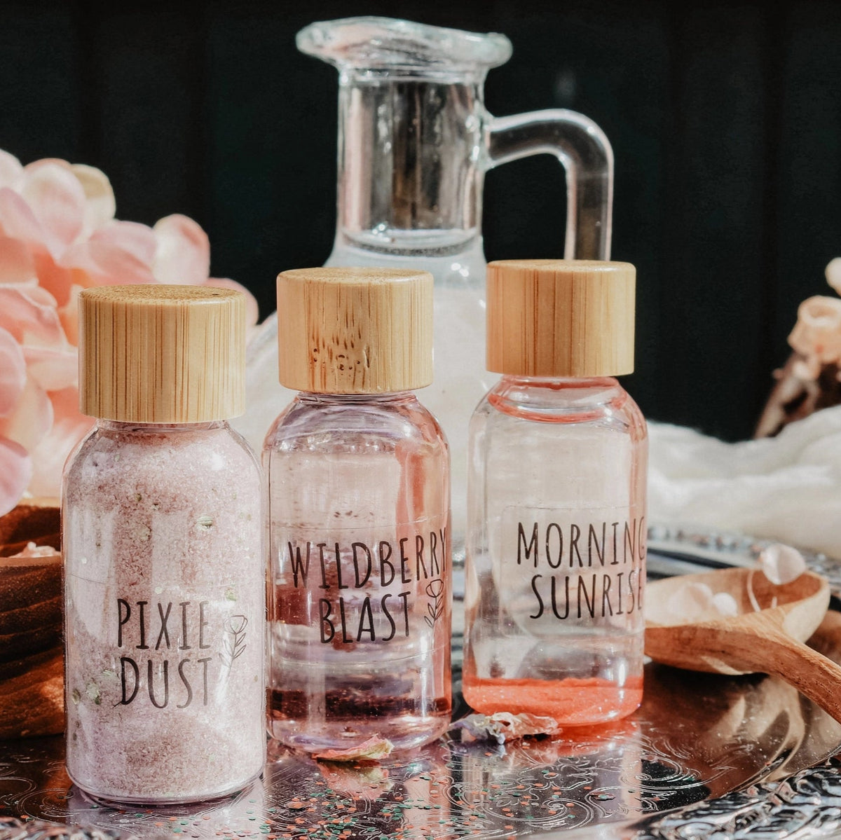 Three small glass bottles with wooden caps on a reflective surface with flowers in the background