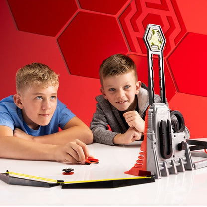 Two boys are playing with a toy sports launcher on a white table against a red geometric background. The launcher is gray and red with a yellow target on top, and the boys are interacting with the toy.