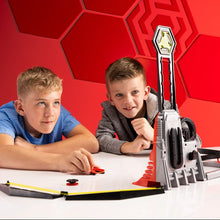 Load image into Gallery viewer, Two boys are playing with a toy sports launcher on a white table against a red geometric background. The launcher is gray and red with a yellow target on top, and the boys are interacting with the toy.
