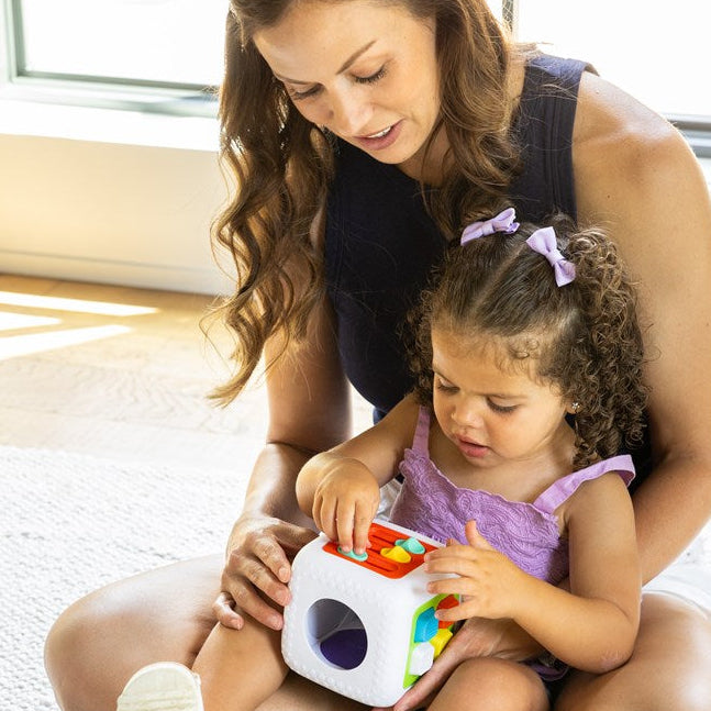 A woman sitting on the floor with a young child holding a white activity cube toy with colorful shapes and holes, in a bright indoor room.