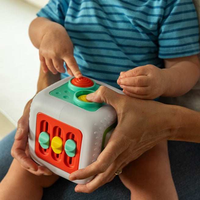 A baby in a blue striped shirt sits on an adult's lap, interacting with a white, red, green, and yellow activity cube toy. The adult's hands are holding the toy and supporting the baby.