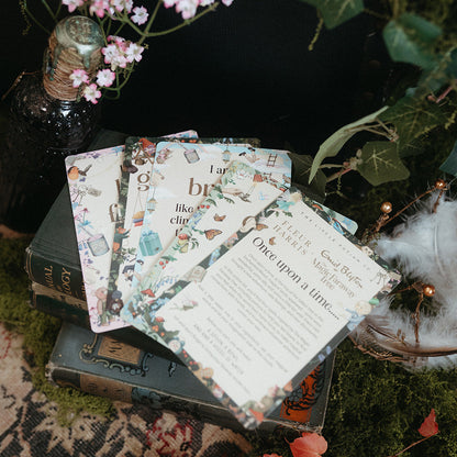 A styled flat-lay of printed cards or invitation sheets with vintage-style illustrations, arranged on stacked books surrounded by moss, flowers, a dark bottle, and small lights.