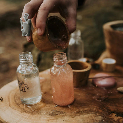 Hand pouring a liquid from a wooden container into two small bottles on a wooden surface.