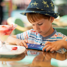 Load image into Gallery viewer, Child wearing a hat using a Purple Cow Travel Game at a dining table with drinks and plates.
