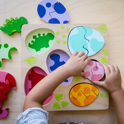 A child's hands interacting with a colorful wooden egg and dino puzzle, with various-sized dinosaur shapes inside egg-shaped compartments.